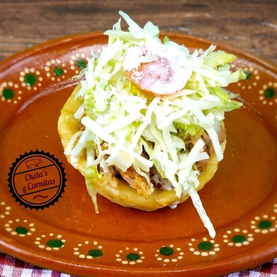 A Chela's y Carnitas sope with shredded meat, lettuce, cream, and tomato on a decorative plate.